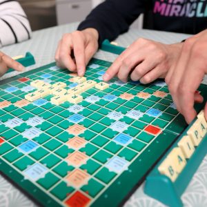 Lähikuva henkilöiden käsistä pelaamassa sanapeli-Scrabblea. Pelilaudalla kirjoitettuna sanat tahto, rauhat ja kuha.