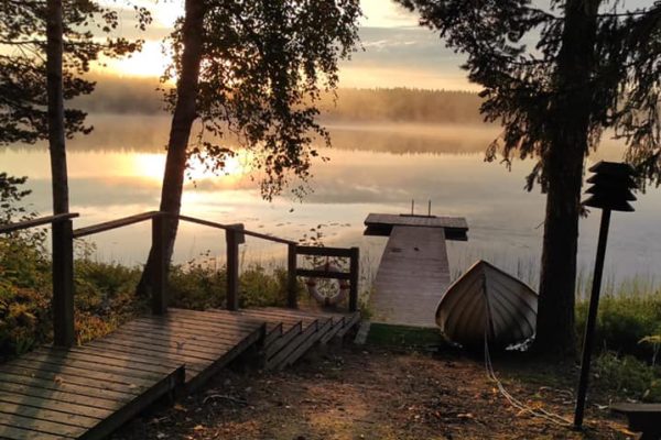 Näkymä Mikkeli-yhteisön rannalta, puiden lomasta usvaisen järvenselän takaa nouseva aurinko. Rannassa laituri ja soutuvene.