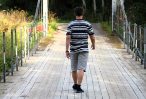 Mies selkä kameraa kohden kävelemässä A-kodin siltaa pitkin eteenpäin. Edessä valkoinen valossa kylpevä portti.