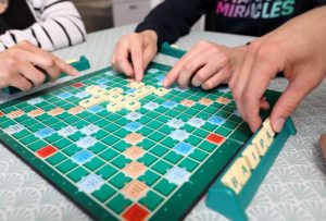 Lähikuva henkilöiden käsistä pelaamassa sanapeli-Scrabblea. Pelilaudalla kirjoitettuna sanat tahto, rauhat ja kuha.